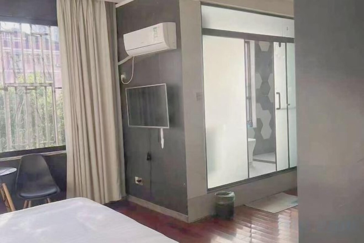 A hotel room with a bed, a TV mounted on the wall, a sliding glass door leading to a bathroom, and a window with curtains.
