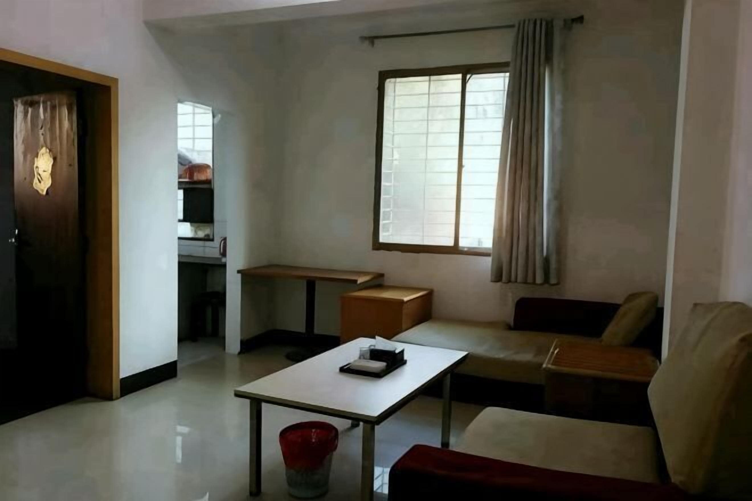 A living room with a sofa, a table, a red bucket, a window with curtains, and a door.