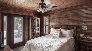 Een slaapkamer in een houten blokhut met een bed, een plafondventilator, een houten dressoir en een boekenplank.