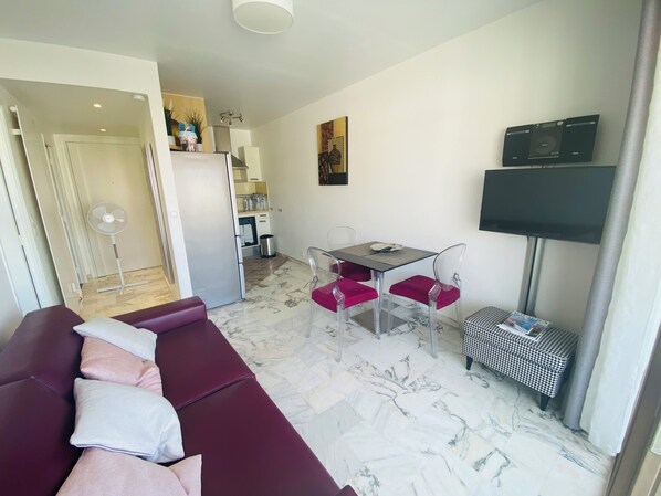 Ein modernes Wohnzimmer mit einer Couch, einem Esstisch mit Stühlen, einem Fernseher und einer Küchenzeile im Hintergrund.