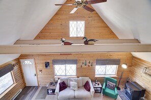 Um quarto acolhedor no estilo loft, com teto de madeira, um sofá branco, uma poltrona verde, lareira e uma janela com vista.