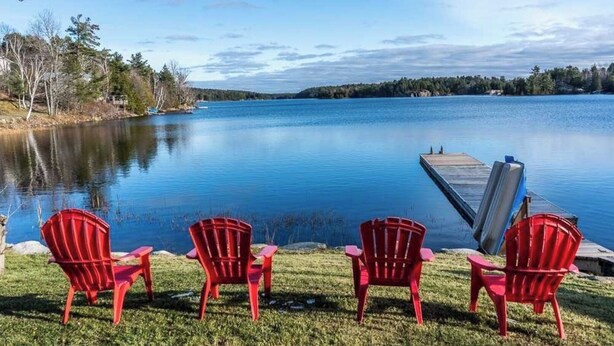 A serene lakeside view with three red Adirondack chairs facing the water, a dock extending into the lake, and a forested shoreline in the background. - Waterfront Cottage with HotTub! Next to OFSC trails (French River)