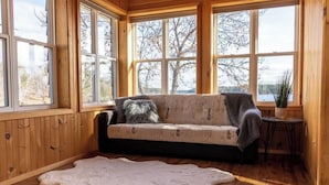 A cozy room with a wooden interior, a sofa with a patterned cover, a large fur rug, and a view of trees through large windows. - Waterfront Cottage with HotTub! Next to OFSC trails (French River)