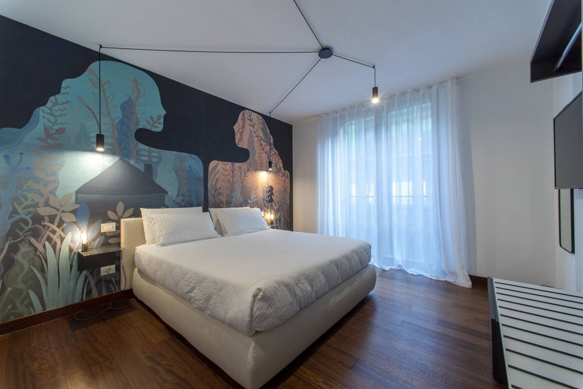 A modern bedroom with a large bed, a mural on the wall, a ceiling light, and a window with curtains.