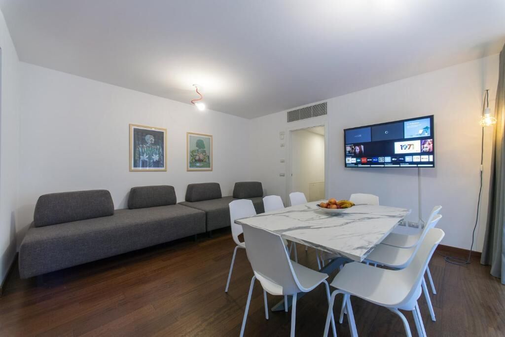 A modern living room with a sofa, a dining table with chairs, a flat-screen TV mounted on the wall, and framed artwork on the wall.
