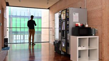 A modern hotel lobby with a large glass door leading to a green outdoor area, a vending machine, and a microwave on a white cabinet.