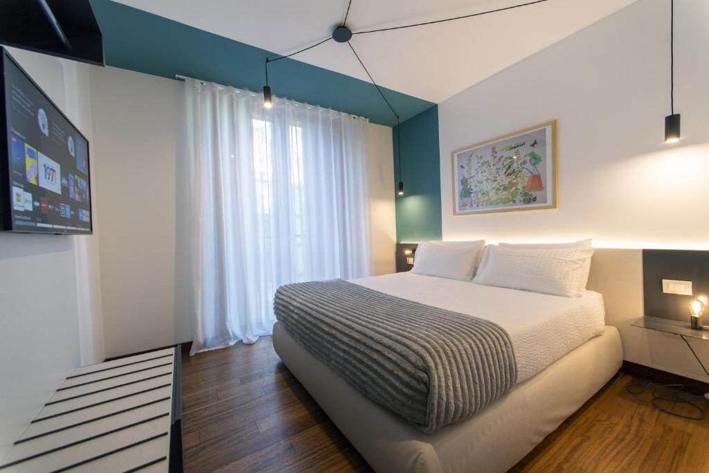 Una habitación de hotel moderna con una cama grande, un televisor de pantalla plana, una ventana con cortinas blancas y un cuadro colgado en la pared.