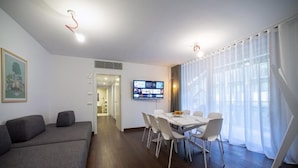 A modern living room with a grey sofa, a dining table with chairs, a flat-screen TV mounted on the wall, and a large window with sheer curtains. - Habitat Apartments (Trento)