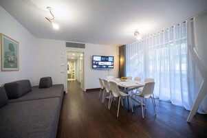 A modern living room with a grey sofa, a dining table with chairs, a flat-screen TV mounted on the wall, and a large window with sheer curtains.