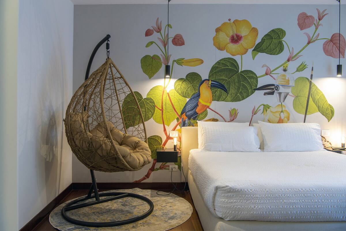 A bedroom with a hanging chair, a bed with white bedding, and a wall mural of tropical flowers and a toucan.