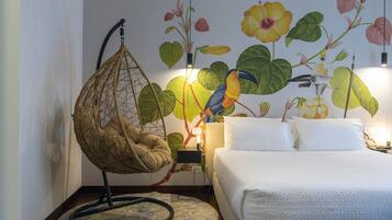 Quarto com uma cadeira suspensa, cama com roupa de cama branca e um mural de flores tropicais e um tucano.