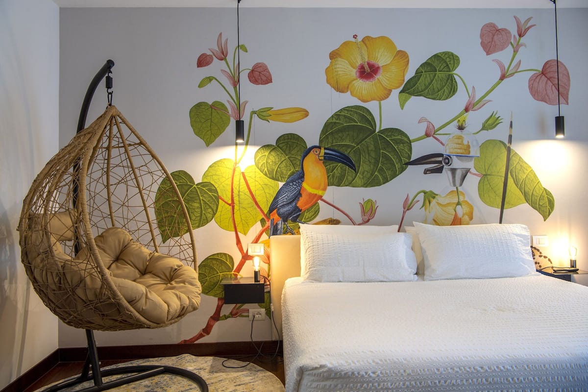 A bedroom with a hanging chair, a bed with white bedding, and a mural of flowers and a bird.