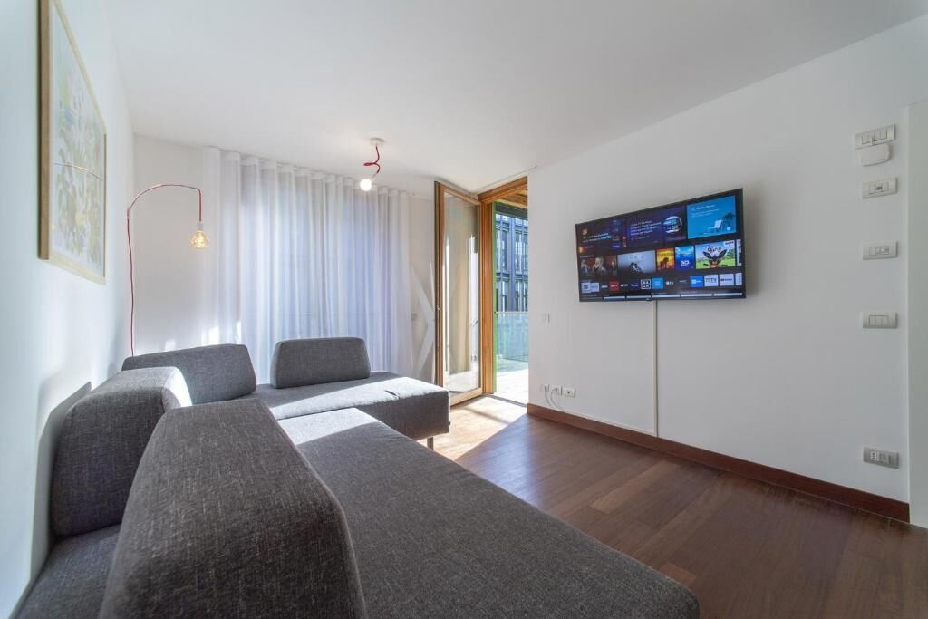 Ein modernes Wohnzimmer mit einem an der Wand befestigten Flachbildfernseher, grauen Sofas, einer Stehlampe und einer Schiebetür zu einem Außenbereich.