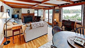 A cozy living room with a fireplace, wooden beams, a sofa, a television, and a view of a lake. - Nest On Mount Desert Isle w Beach - 8 Mi to Acadia (Mt Desert)
