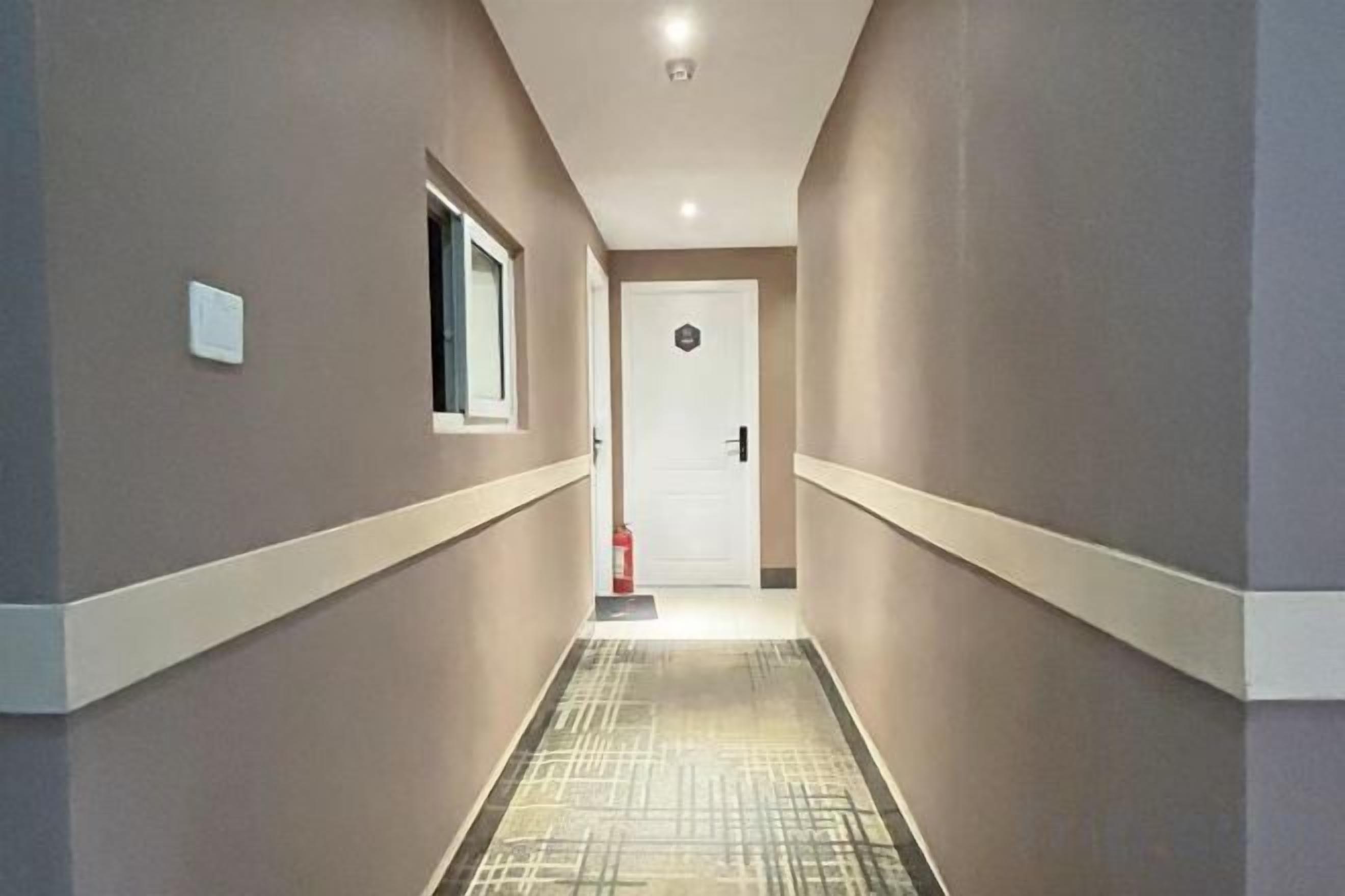 Un pasillo con alfombra estampada, una puerta blanca al final y una pequeña ventana en la pared izquierda.