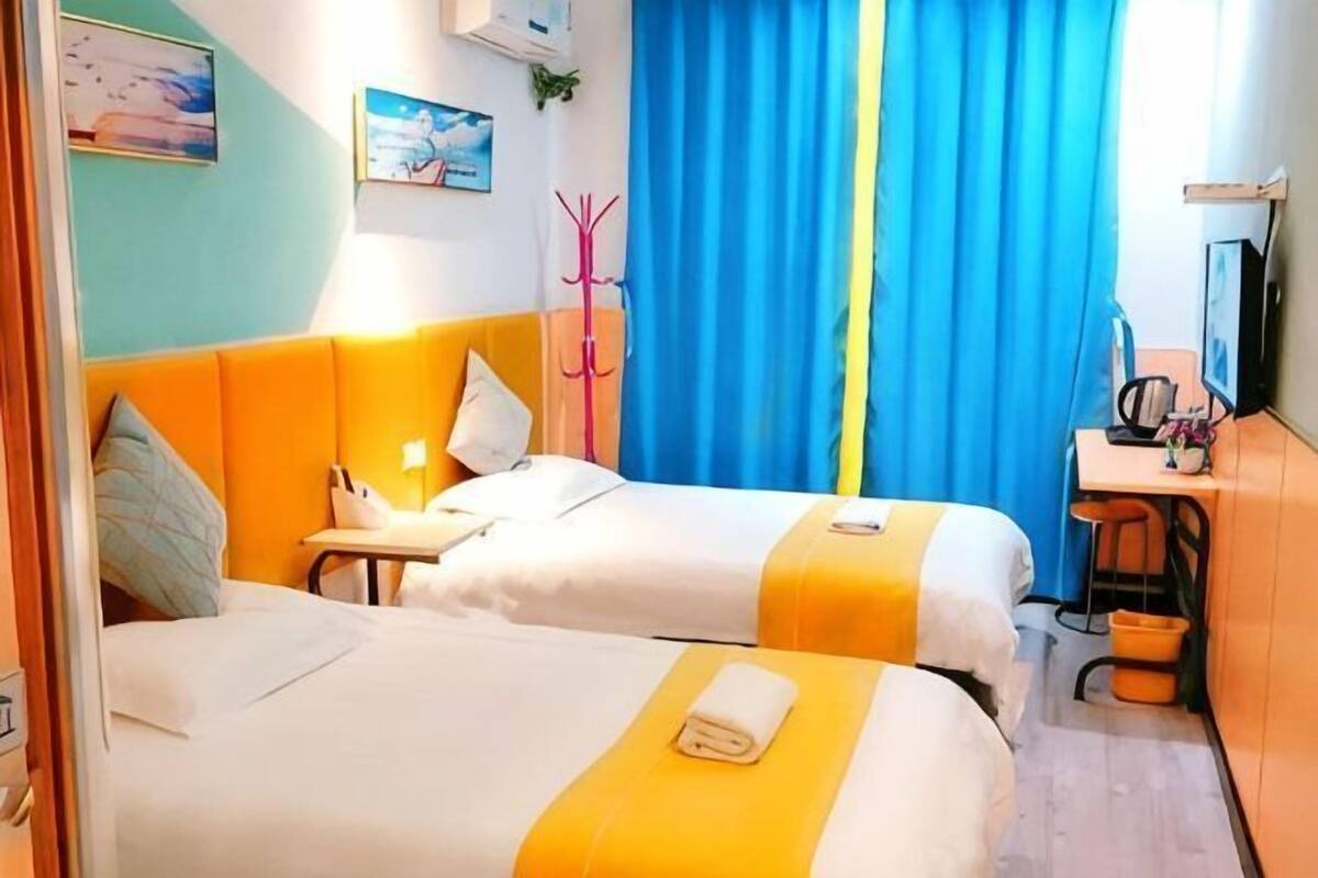 A hotel room with two beds, a desk, a chair, a coat rack, and a window with blue curtains.