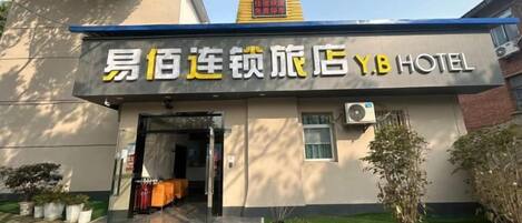 A hotel entrance with a clear doorway, a signboard, and a small garden area with potted plants.