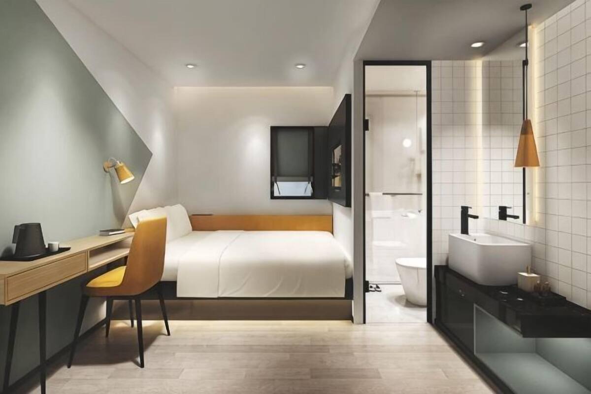 A modern hotel room with a bed, desk, chair, and a bathroom with a sink and toilet.