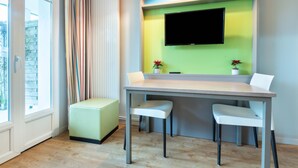 A modern room with a wooden floor, a table with two chairs, a TV mounted on a green wall, a window with curtains, and a small plant on the table. - AR MOR 603 - One Bedroom Apartment, Sleeps 4 (Courseulles-sur-Mer)