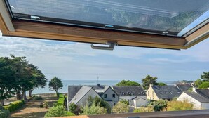 A view through a window with a wooden frame and a mesh screen, overlooking a coastal area with houses and greenery. - Côté Mer - Maison familiale accès privé à la plage (Sarzeau)