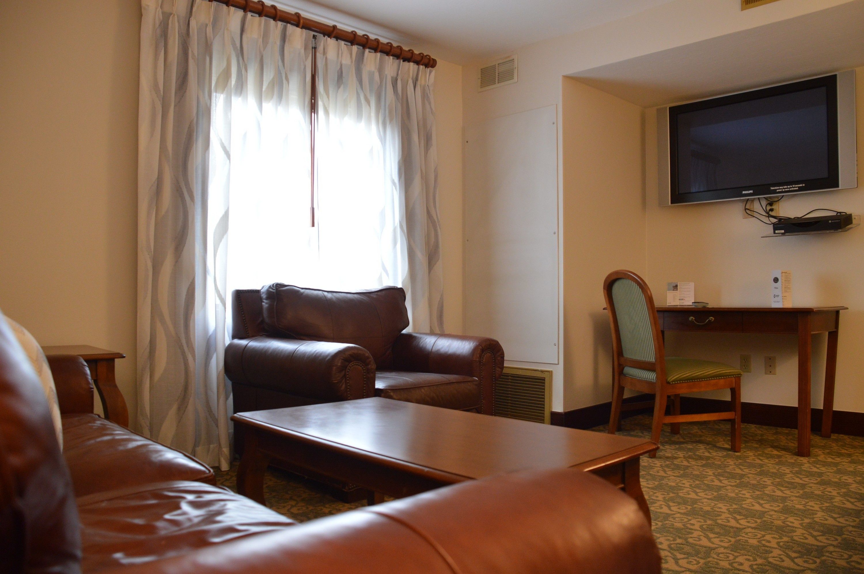 A hotel room with a brown leather sofa, a wooden coffee table, a wooden desk with a chair, and a flat-screen TV mounted on the wall.