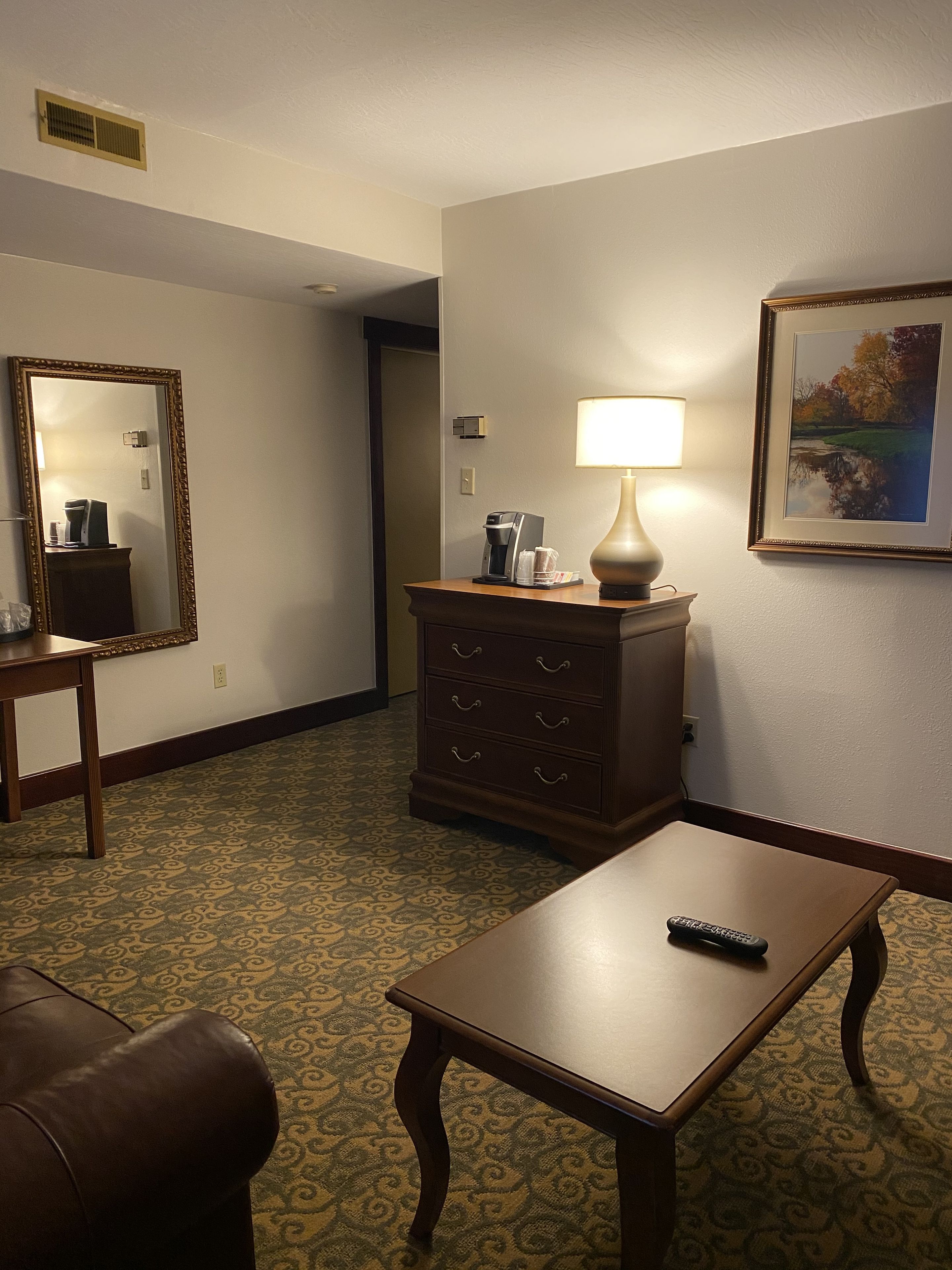 Ein Hotelzimmer mit einem gemusterten Teppich, einer Holzkombination mit Lampe, einem gerahmten Bild an der Wand und einer Fernbedienung auf einem Tisch.