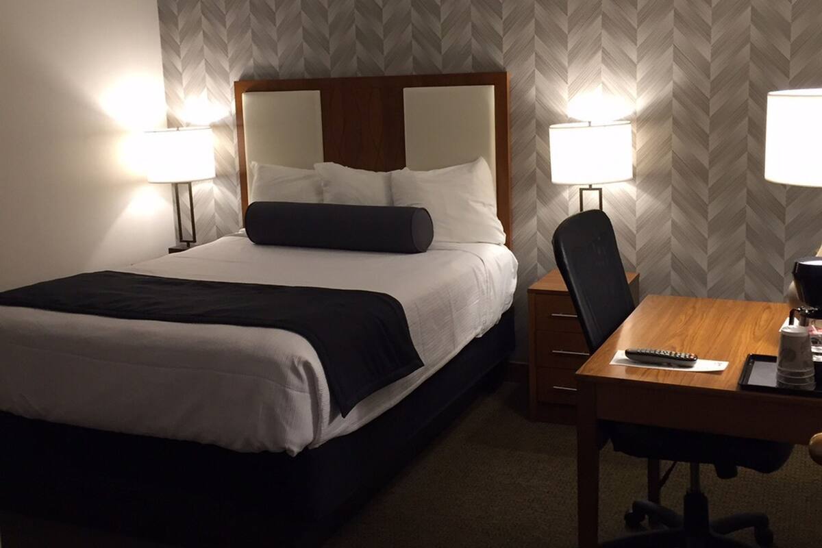 A hotel room with a bed, two bedside lamps, a wooden desk, and a chair.