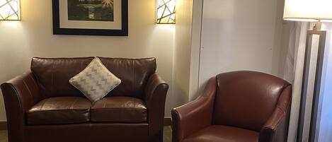 A brown leather sofa with a patterned pillow, a brown leather armchair, and a floor lamp in a room with a patterned carpet.