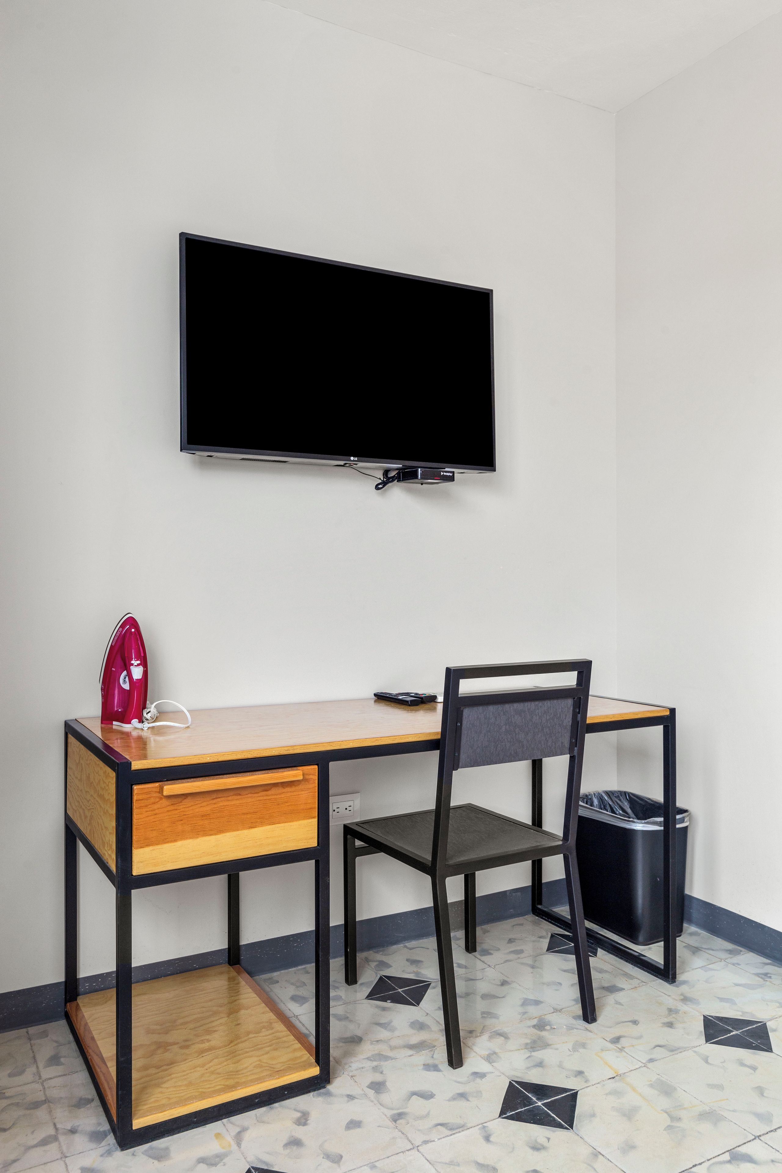 Una habitación pequeña y moderna con un escritorio de madera, una silla, un televisor montado en la pared y un cesto de basura.