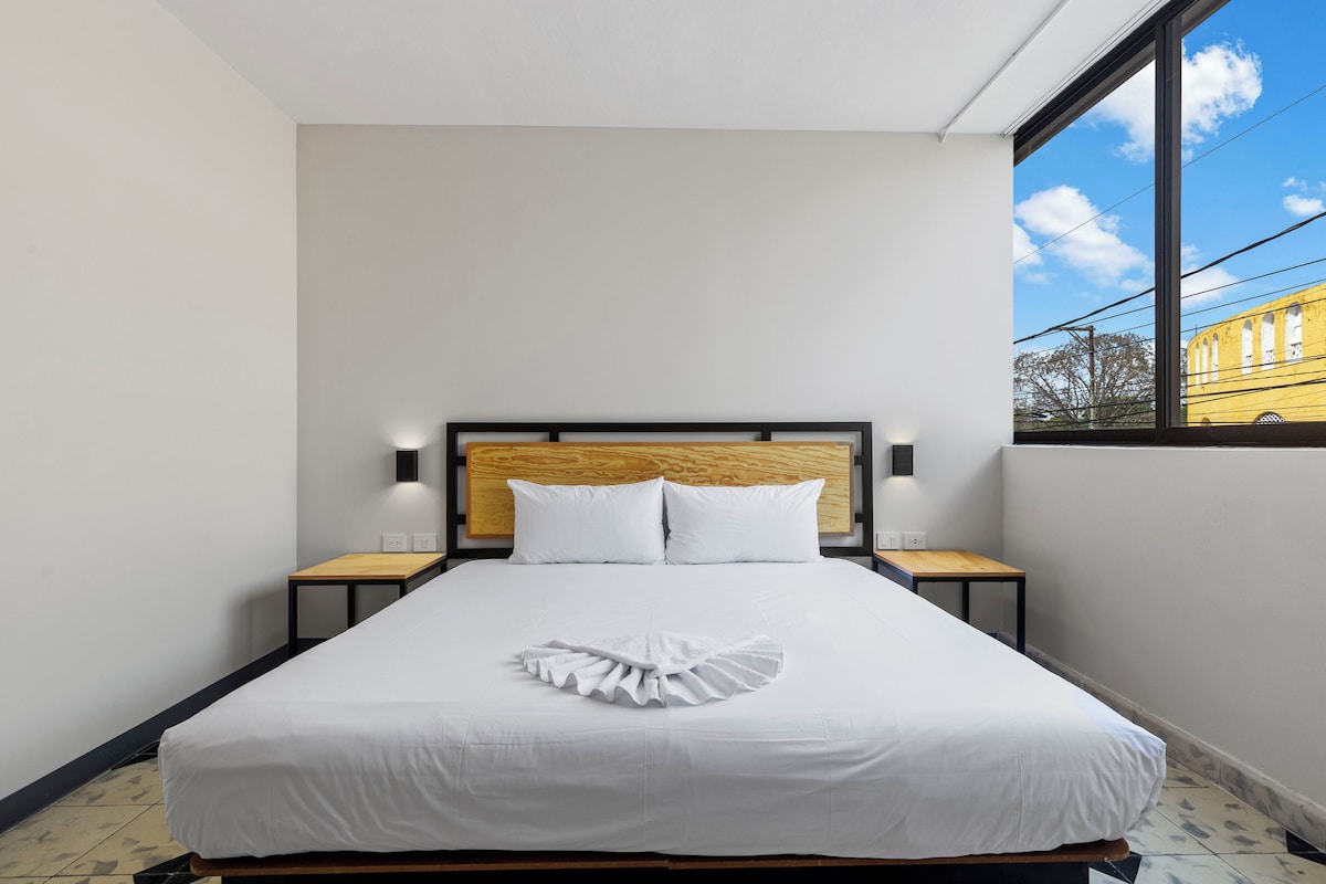 A hotel room with a large bed, a wooden headboard, two bedside tables, and a view of a building through the window.