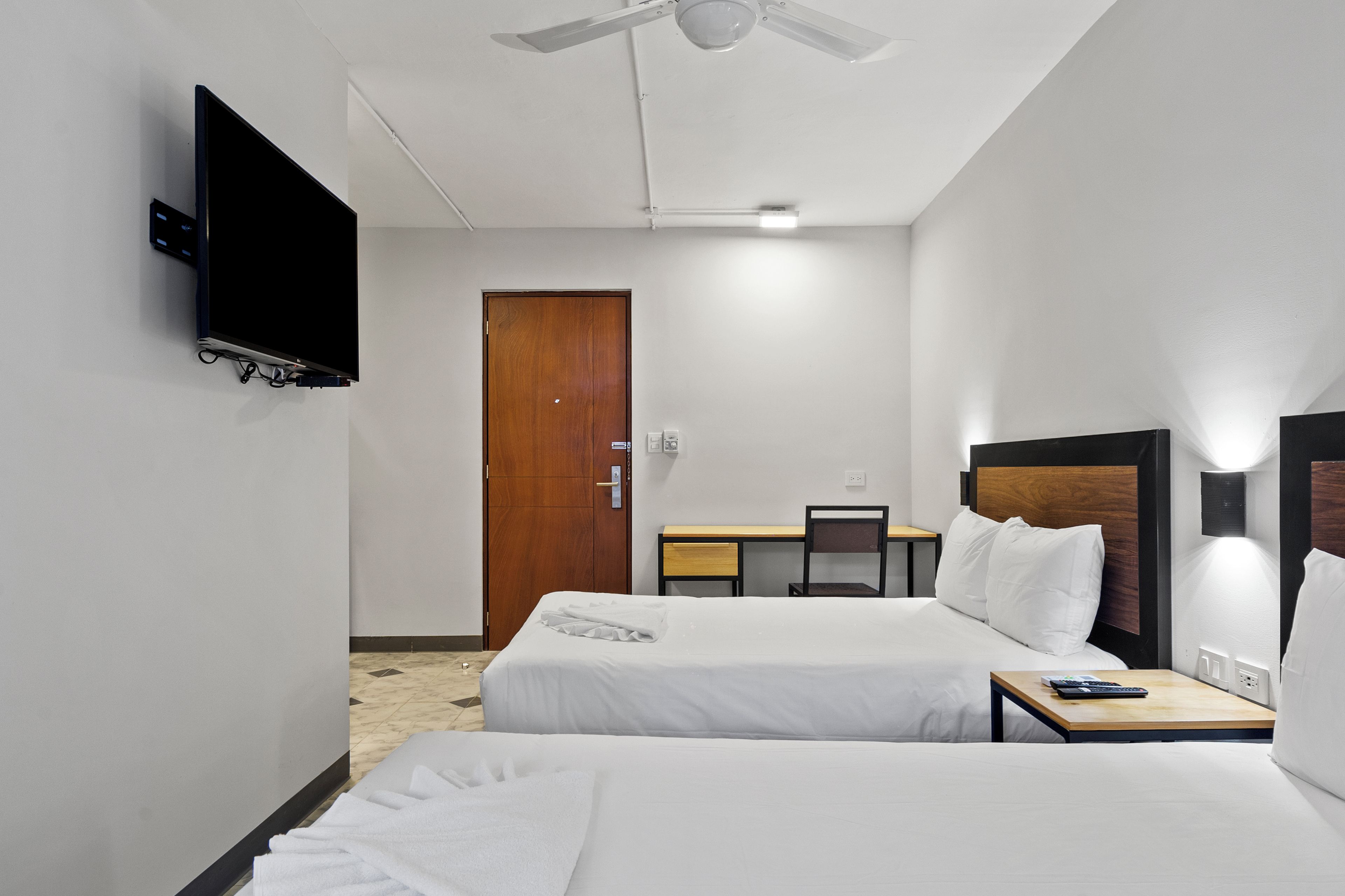 A hotel room with two beds, a desk, a chair, a TV mounted on the wall, and a ceiling fan.