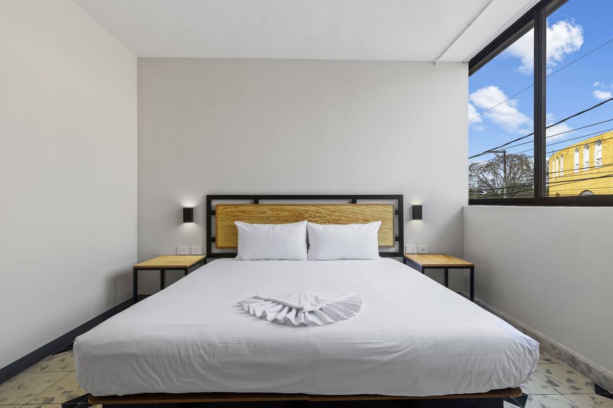 A neatly made bed with white linens and a decorative pillow, a wooden headboard, bedside tables with lamps, and a view of a building through a large window.