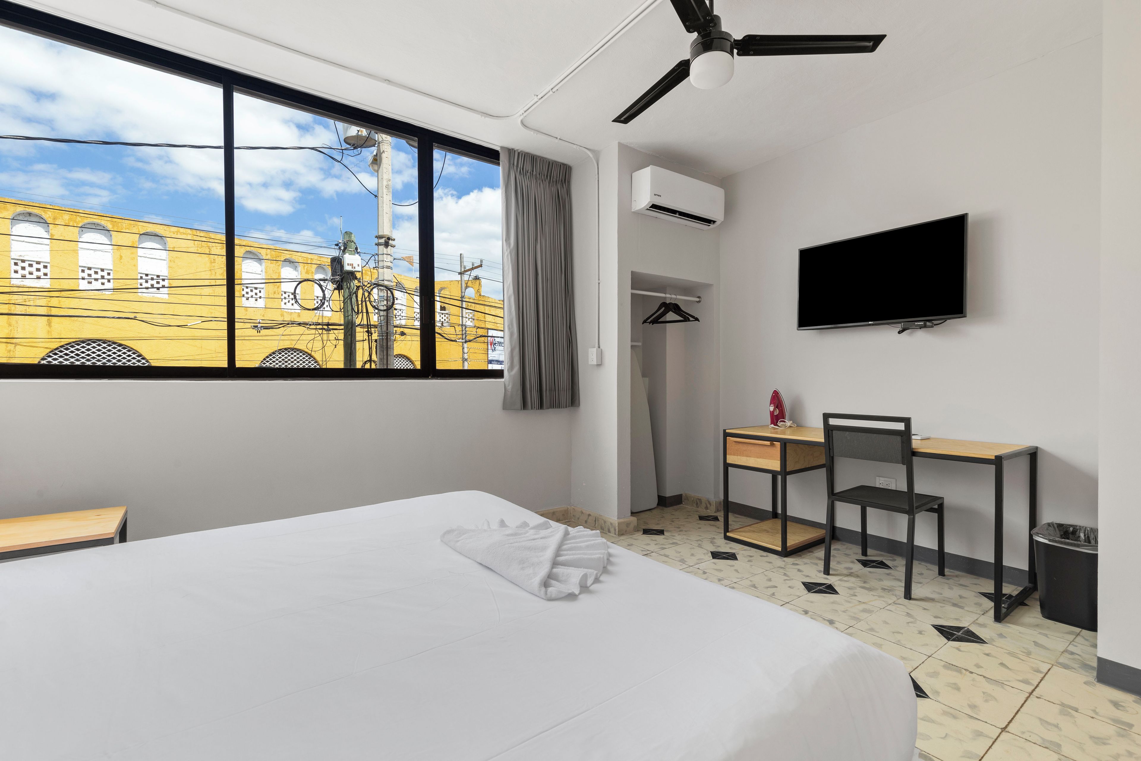 A hotel room with a bed, a desk, a chair, a TV, and a view of a yellow building.