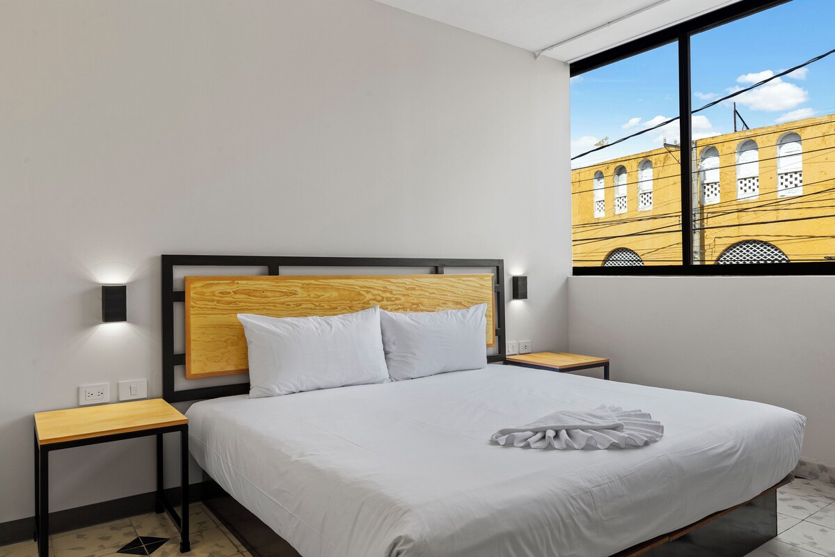 Une chambre d’hôtel moderne avec un grand lit, une tête de lit en bois, du linge de lit blanc et une vue sur un immeuble jaune par la fenêtre.