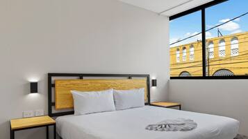 Habitación de hotel con cama, dos mesitas de noche, ventana con vista a un edificio amarillo y cabecero de madera.