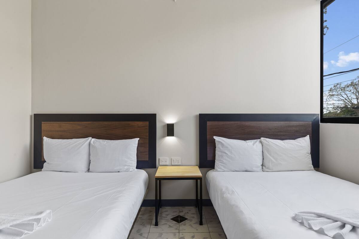A hotel room with two single beds, a small table, and a nightstand.