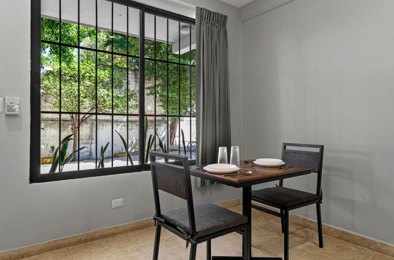 Un comedor con una mesa dispuesta para dos personas, sillas, una ventana con vistas a zonas verdes y una cortina.
