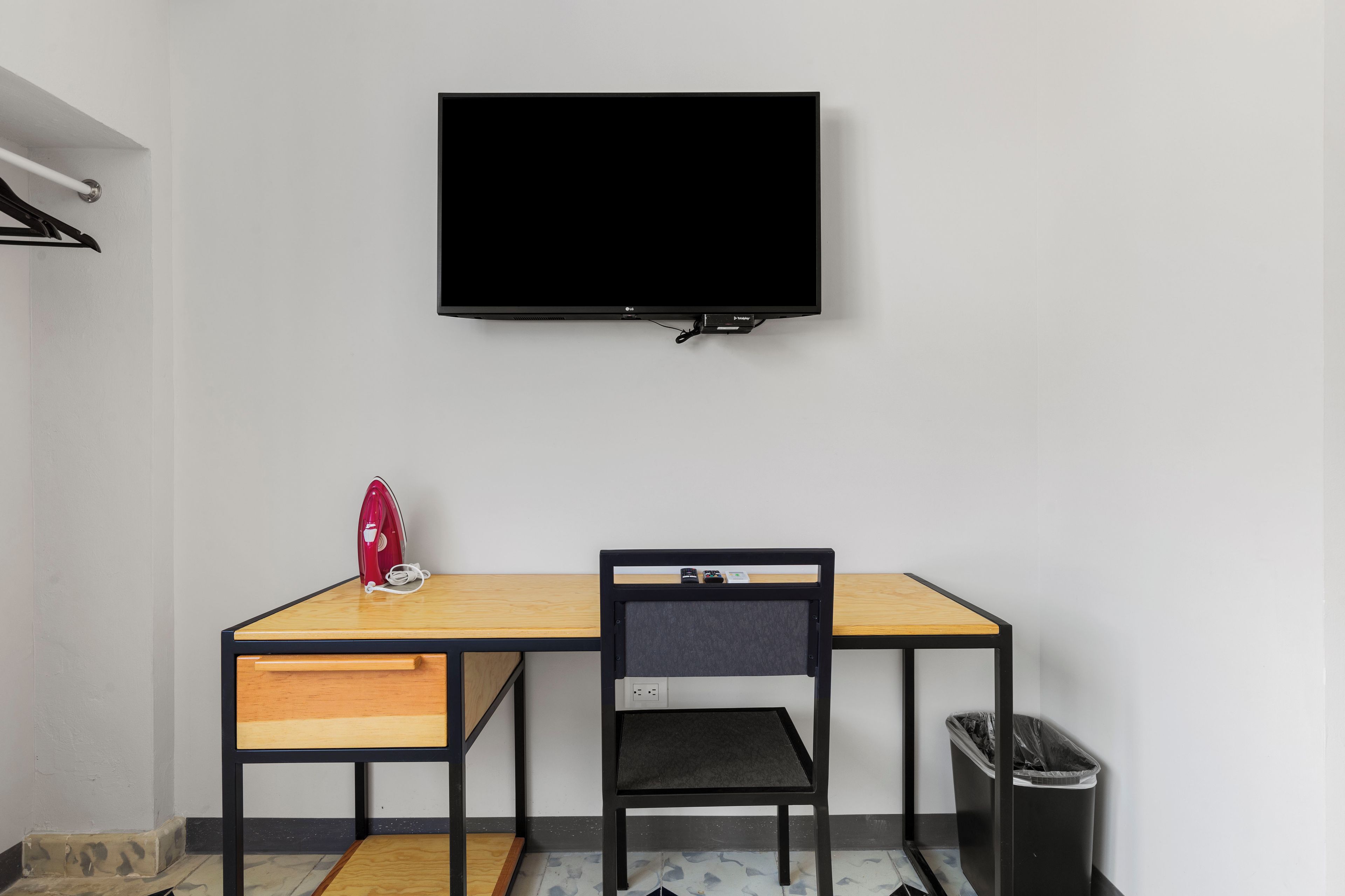 A room with a TV mounted on the wall, a desk with a chair, a red iron, and a trash bin.