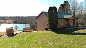 A cabin with a deck overlooking a lake, surrounded by trees and a grassy area. - Quiet, Cozy Paoli Cabin Overlooking a Nice Pond (Paoli)