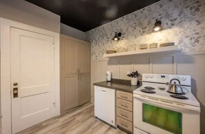 Una cucina con piano cottura bianco, lavastoviglie e un piano di lavoro con un bollitore. Si vedono armadietti e una porta.