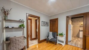 Una habitación con suelo de madera, un sillón tapizado, una mesita con una planta y una puerta que da a otra habitación.