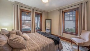 Un dormitorio con una cama grande, dos ventanas, una silla y un espejo.