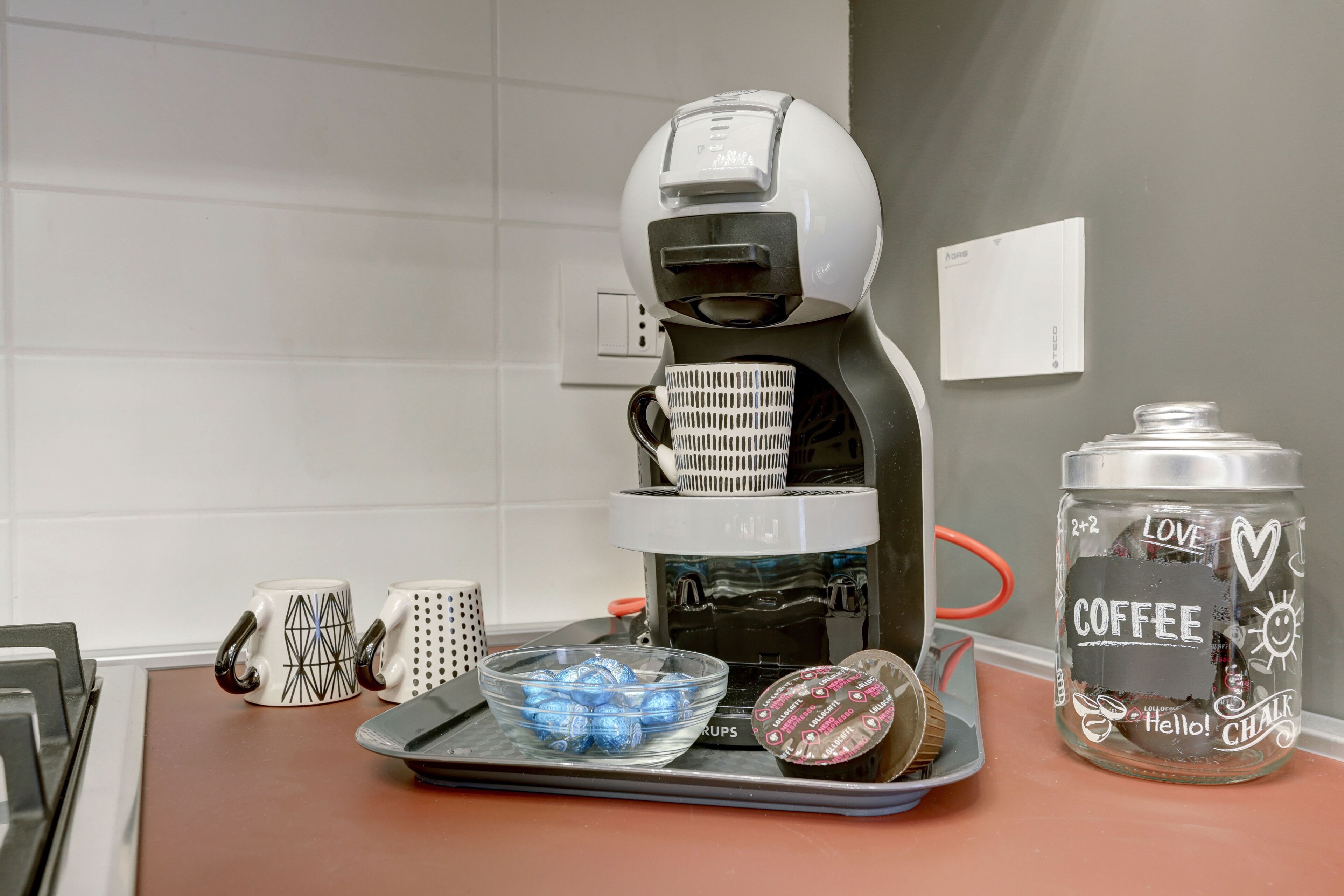 Uma cafeteira sobre uma bancada de cozinha, com um pote de café e duas chávenas.