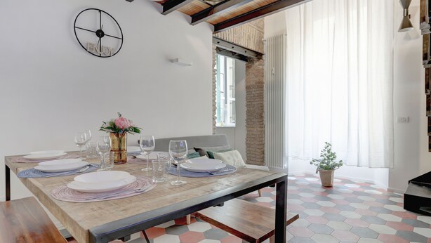 Un comedor con una mesa de madera dispuesta para cuatro, un reloj en la pared y una pared de ladrillo detrás de la ventana.