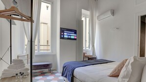 Una habitación de hotel con una cama, un televisor montado en la pared, un ventanal con cortinas y una mesita de noche con toallas y una botella.