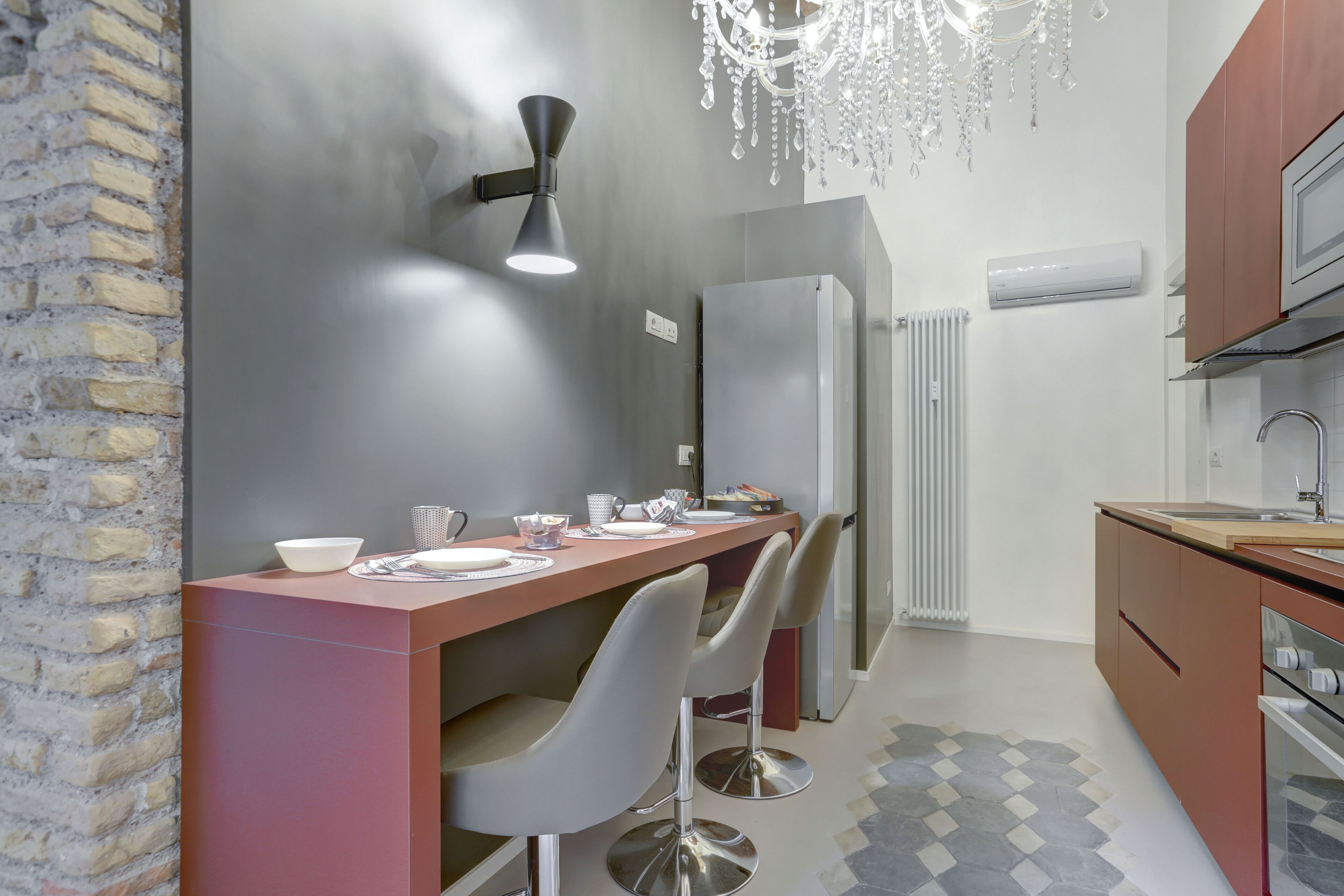 A modern kitchen with a long countertop, two bar stools, a chandelier, and a refrigerator.