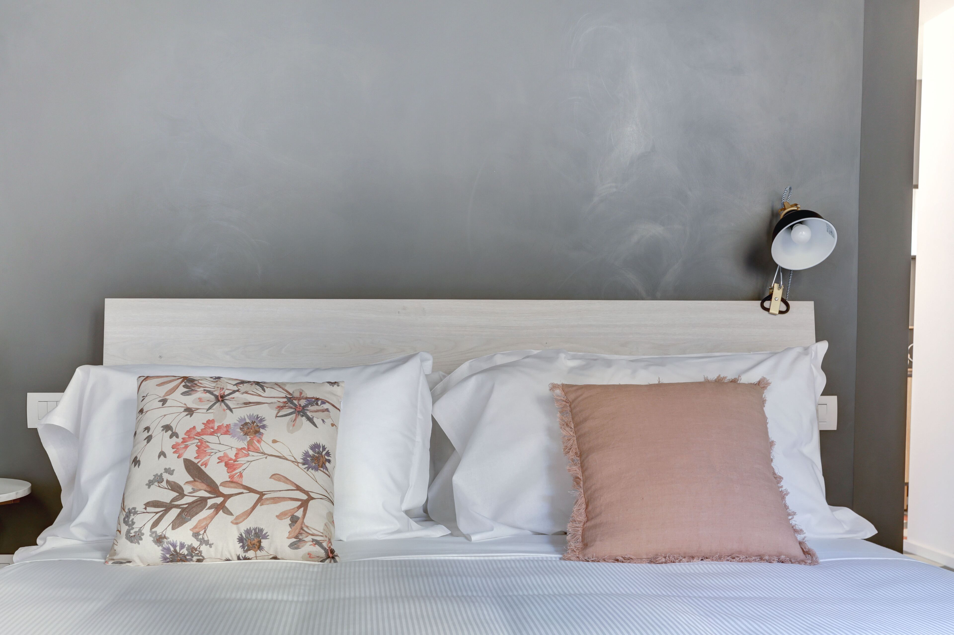 A neatly made bed with floral and solid pillows, a bedside table, and a wall-mounted lamp.