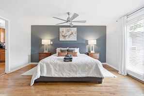Un dormitorio con una cama, dos mesitas de noche, un ventilador de techo, un cuadro en la pared y una ventana con persianas.