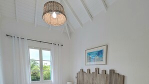 Quarto com uma cama grande, janela com cortinas, luminária pendente, mesa de cabeceira com abajur e quadro na parede.