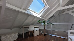 Quarto sótão amplo com pé direito alto, claraboia, piso de madeira, escrivaninha, cadeira, planta em vaso e uma cama.